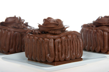 Three Square chocolate cake cupcakes on a chocolate candy bar frosted with frosting rose on top, sitting on square white plates isolated on white background