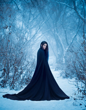 fairy tale girl a demon walks alone in winter forest. Gothic Woman wearing  long black traveling cloak dress with train. Cold nature white snow blue tree wood. Mystery Lady witch back rear view