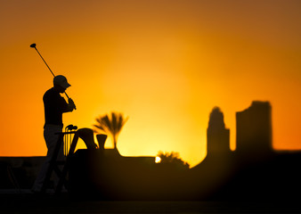 Obraz premium Golf player silhouette on a colorful sunset holding a golf stick. Buildings, sun and palm tree on the background.