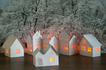 Close up of handmade small white cardboard houses with illuminated windows on snowy trees...