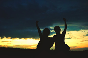 two female silhouettes sitting in front of a sunset sky with their arms raised