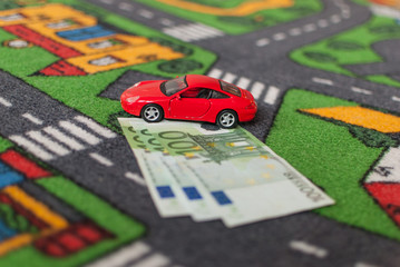Red toy car parked on three 100 euro bills on street themed carpet, transportation costs concept