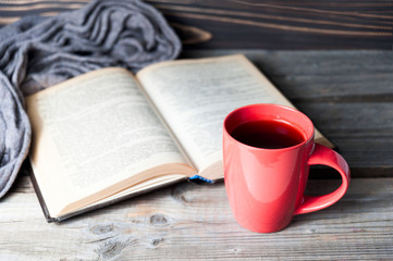 selective focus photo of grey cozy knitted scarf with cup of coffee or tea and open book on a wooden table.