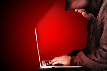 Male computer hacker wearing a hooded top leaning over a laptop in the dark. The screen light illuminates the man with a beard performing illegal activities. Black copy space on the left