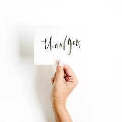 Minimal pale composition with girl's hand holding card with quote Thank You written in calligraphic style on paper on white background. Flat lay, top view
