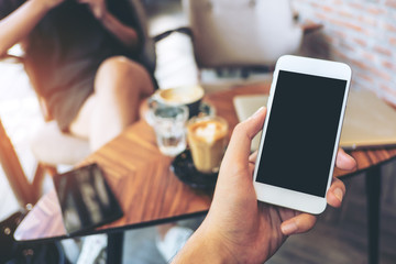 Mockup image of hand holding white mobile phone with blank black screen in vintage cafe and blur asian woman 