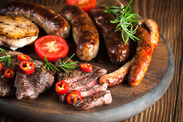 Close-up photo of mixed grilled meat platter. Beef, pork, poultry, sausages, grilled garlic, chili pepper, red tomatoes on wooden rustic background.