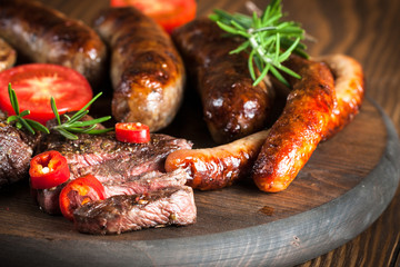 Close-up photo of mixed grilled meat platter. Beef, pork, poultry, sausages, grilled garlic, chili pepper, red tomatoes on wooden rustic background.