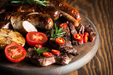 Close-up photo of mixed grilled meat platter. Beef, pork, poultry, sausages, grilled garlic, chili pepper, red tomatoes on wooden rustic background.