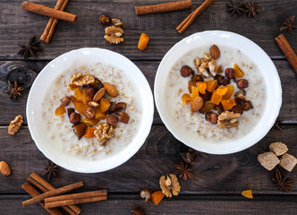 Fototapeta premium Two plates of homemade oatmeal porridge with dried dates, apricots, almonds, hazelnuts, walnuts on old dark grey rustic wooden table. Hot and healthy food for Breakfast. Cinnamon and for decoration