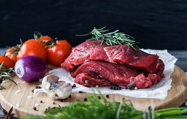 Raw meat beef steaks slices ready for roasting on a wooden cutting board with fresh organic vegetables, onion, garlic, spices, peppercorns, rosemary, sea salt, cherry tomatoes, dill and herbs