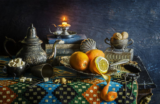Classic still life with fresh oranges in silver plate placed with illuminated candle,old books, vintage kettle,nuts, sugar on ornate cloth..