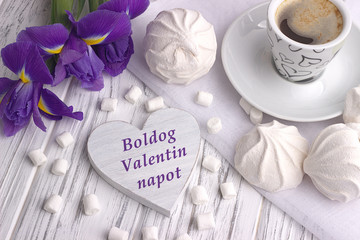 Still life with cup of coffe marshmallow zephyr iris flowers heart sign with lettering Happy Valentines Day in Hungurian on white wooden background.