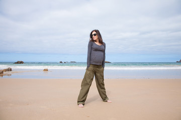 brunette brown hair pregnant woman with grey jersey green trousers and black sunglasses standing in sand beach ocean in Asturias Spain

