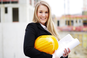 Junge Frau mit Helm und Plänen vor einer Baustelle