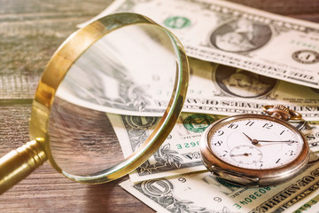 Time is money finance concept with old vintage clocks, dollar bills and magnifying glass on wooden table
