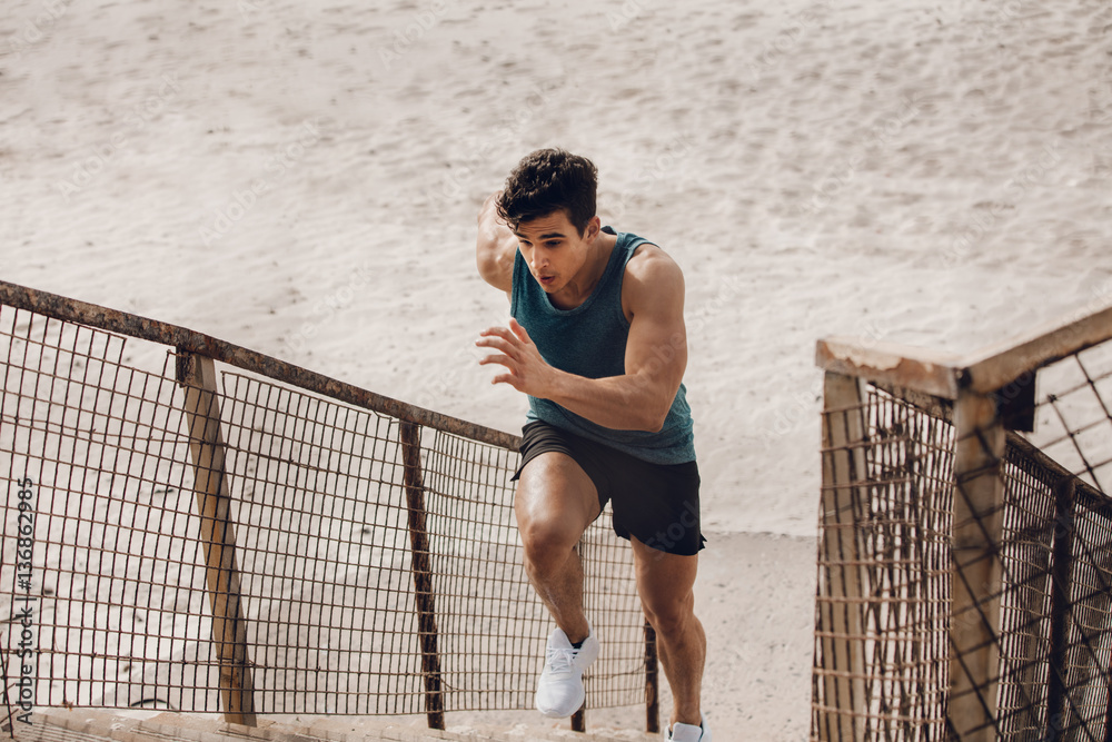 Wall mural Fit young man running up the steps at beach