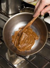Rich thick brown curry paste being mixed and cooked in a metal frying pan, over a metal kitchen hob.