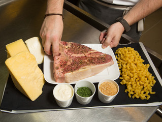 A fresh raw T bone steak meat cut, covered in chopped up pepper corns, on a plate with macaroni noodles, cheese and cream.