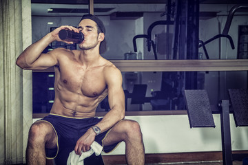 Attractive athletic shirtless young man drinking protein shake from blender in gym while looking at...