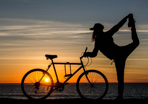 Silhouette of girl with bicycle at sunset time. Young woman is doing yoga near the ocean with bicycle