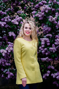 Blonde girl in a yellow coat stands against the backdrop of blooming lilacs in spring