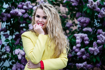 Blonde girl in a yellow coat stands against the backdrop of blooming lilacs in spring