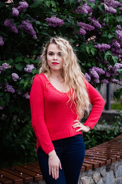 Blonde girl in a yred sweater stands against the backdrop of blooming lilacs in spring