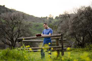 Man reading a book outside in a field leaning against a wooden fence.