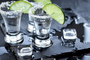 Silver tequila shots with ice and lime on black table background