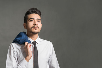 Young attractive man in a white shirt, tie and jacket hanging on his shoulder on a gray background