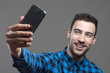 Excited young smiling man taking self portrait with mobile phone over gray studio background. 