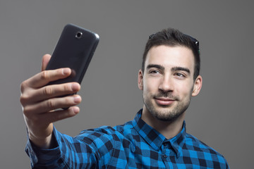 Moody portrait of young confident happy man taking self portrait with mobile phone over gray studio background. 