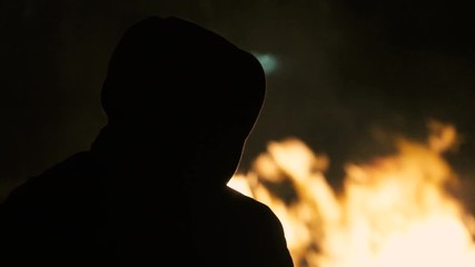 Silhouette of a hooded man staring at a bonfire burning in the night
