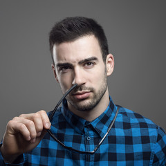 Moody portrait of skeptical young man taking off sunglasses looking at camera dubiously over gray studio background. 