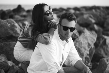 Young Happy fashionable couple resting by the sea on sunset. The man and woman Wearing in romantic sexy summer clothes and  in stylish trendy sunglasses