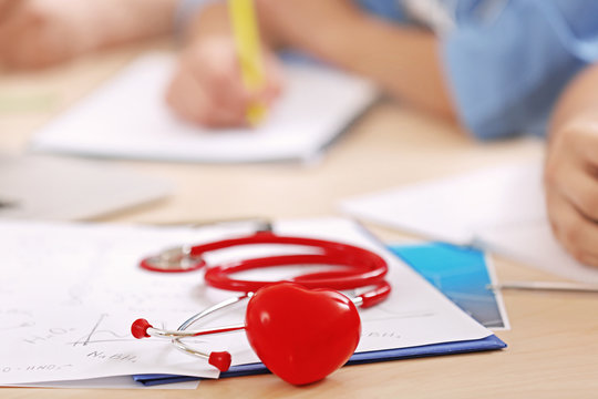 Stethoscope, miniature heart and clipboard on desk and interns on background, closeup
