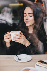 Beautiful young woman enjoying coffee cappuccino with foam near window in a cafe