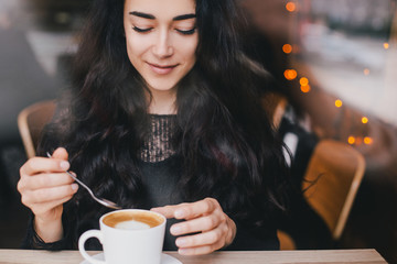 Beautiful young woman enjoying coffee cappuccino with foam near window in a cafe