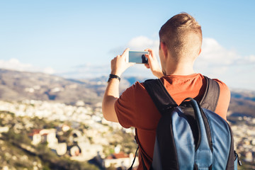 Back view of a young man tourist with backpack taking photo of landscape with smartphone