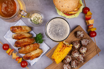 Glasses of beer with chicken wings, burger, meat balls, grilled corn and vegetables. Beer bites. Ale and food still life. View from above, top studio shot