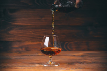 Pouring brandy or cognac from the bottle into the glass against wooden background