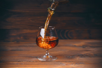 Pouring brandy or cognac from the bottle into the glass against wooden background