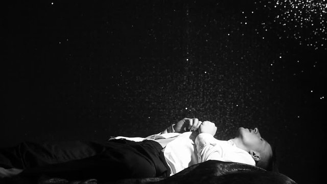 Man lies on a bed and buttoning a white shirt on black wall background at night. Loneliness, depression, fatigue and lack of motivation. Young man dreaming in bed before work