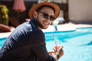 Man in sunglasses and hat drinking cocktail, sitting near pool.
