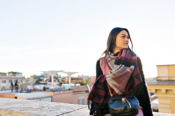 Young woman is sitting on the roof with beautiful view on sunny city. She looks relaxed. She is wearing in dark pullover and bright scarf.