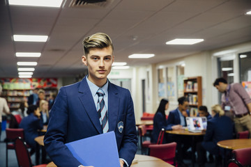 Teen student is posing for the camera in his classroom. He is holding a book and is wearing school uniform.