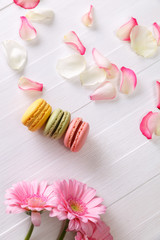 Macaroon cakes with pink rose petals and Gerbera flowers. Different types of macaron. Colorful almond cookies. On white wooden rustic background.