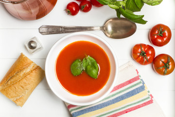Sopa crema de tomate en cuenco blanco con albahaca sobre mesa blanca con pan cuchara y servilleta. Vista superior
