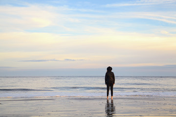 Young woman with long hair standing on wet sand on beach and looking away. Female in black hat relaxing on the sea ocean on sunset. Back view. Peaceful scene, calming waves, pastel cloudy sky, coast.